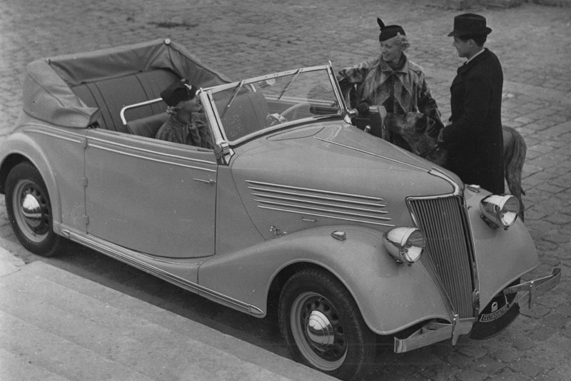 Photo noir et blanc d'un homme et une femme debout à coté d'une voiture des années 30 avec une passagère a bord