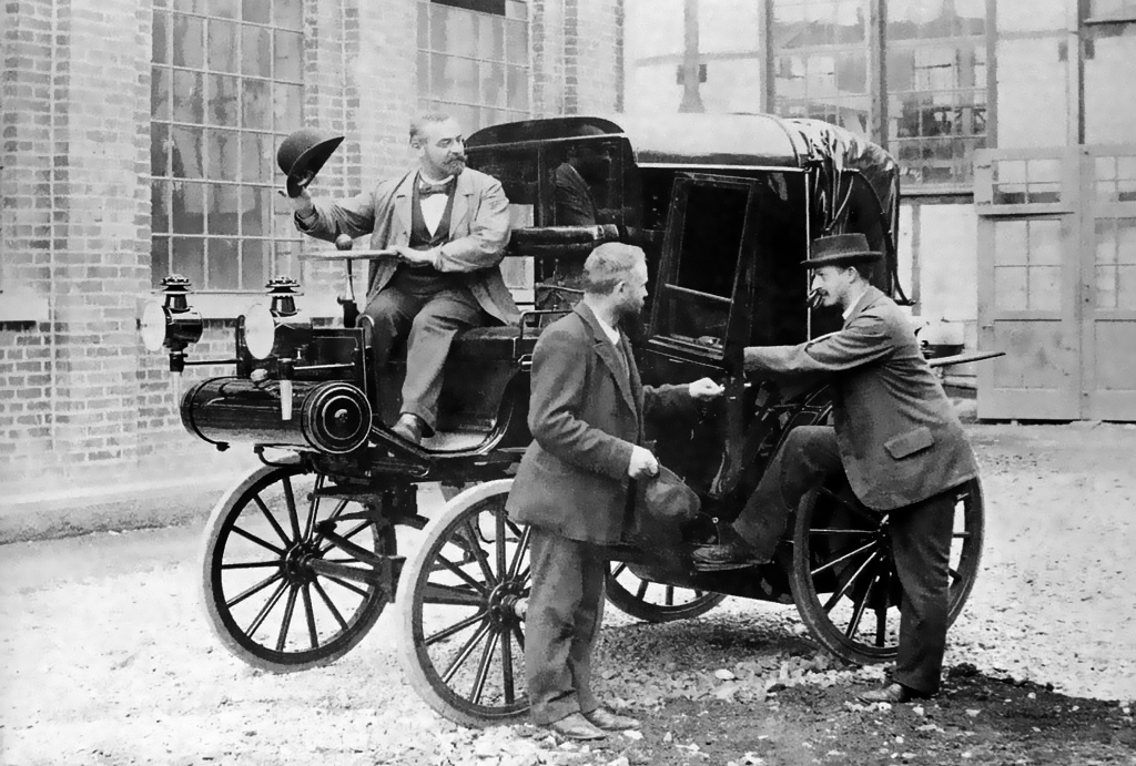 Photo noir et blanc d'une voiture de fin du 19eme siècle avec un chauffeur assis au volant et deux personnes debout a coté réalisant une transaction financière